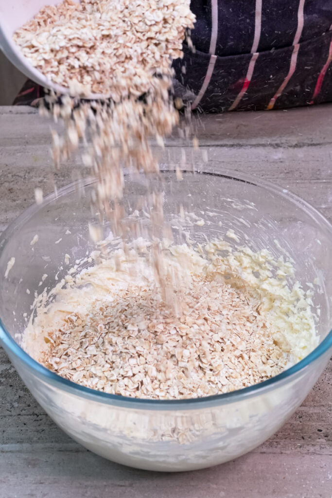 Old-fashioned rolled oats being added to the oatmeal cookie dough in a clear glass bowl