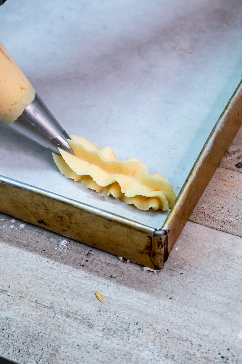 Piping long, fluted lines of churro cookie dough onto a baking sheet lined with parchment paper