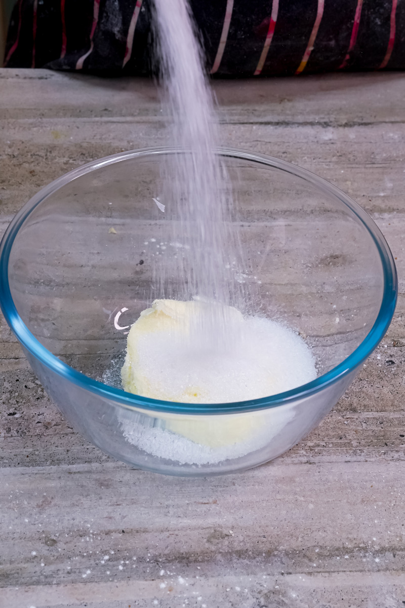 White granulated sugar being poured from a cup into a clear glass bowl with softened butter