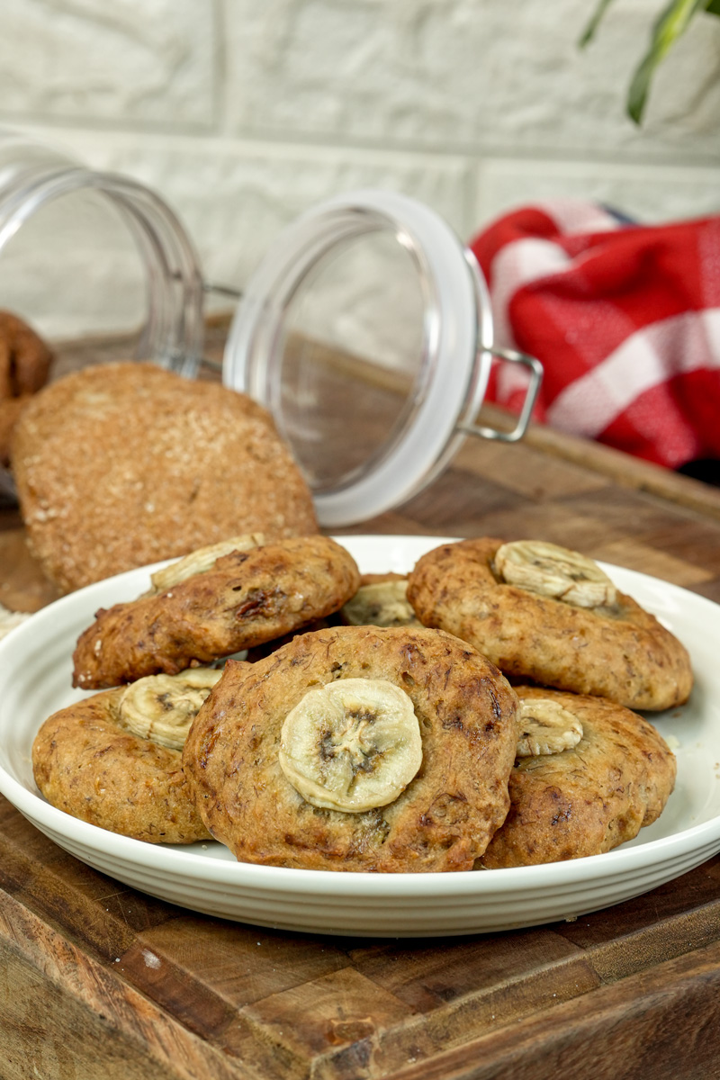 A rustic shot of a plate of banana cookies with a glass jar and a red plaid napkin in the background