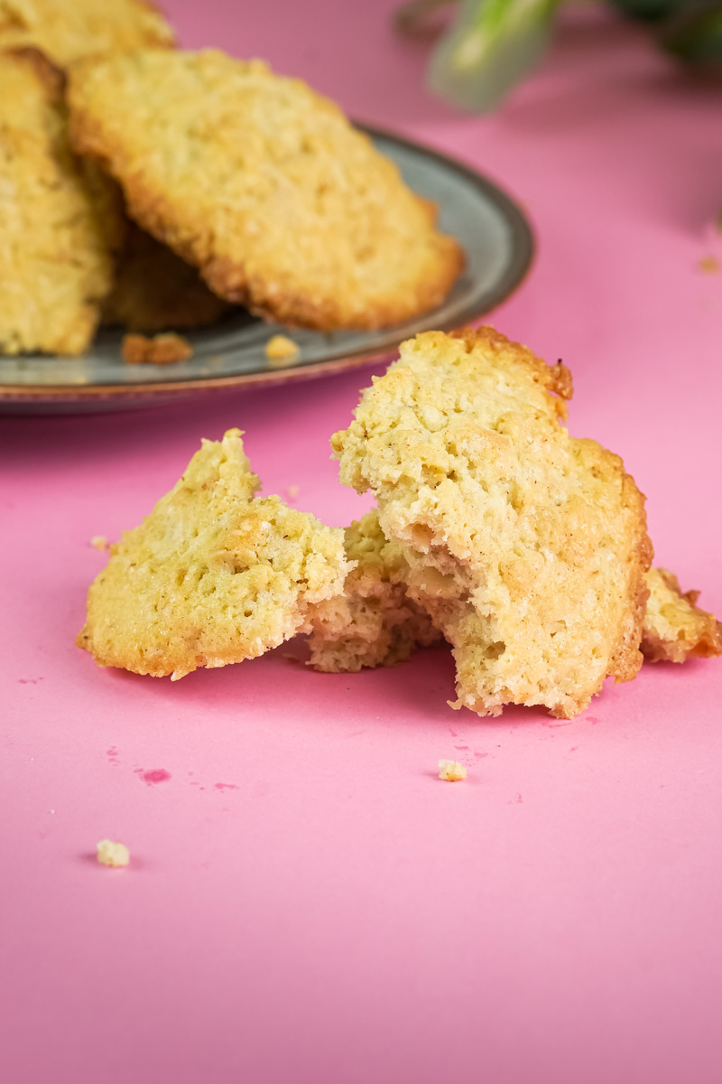 A broken oatmeal cookie sits on a pink surface with crumbs, showing a crunchy exterior and soft interior