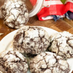 A plate of chocolate crinkle cookies, with a cookie jar and festive red checkered towel in the background
