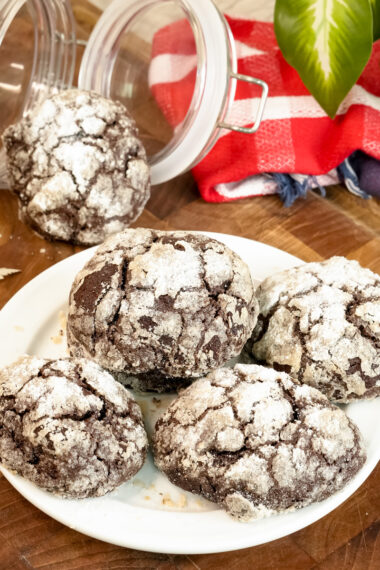 A plate of chocolate crinkle cookies, with a cookie jar and festive red checkered towel in the background