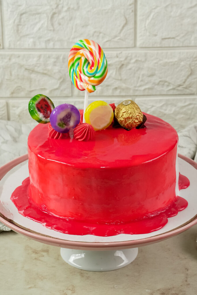 A completed drip cake decorated with a lollipop and candies on a pink cake stand
