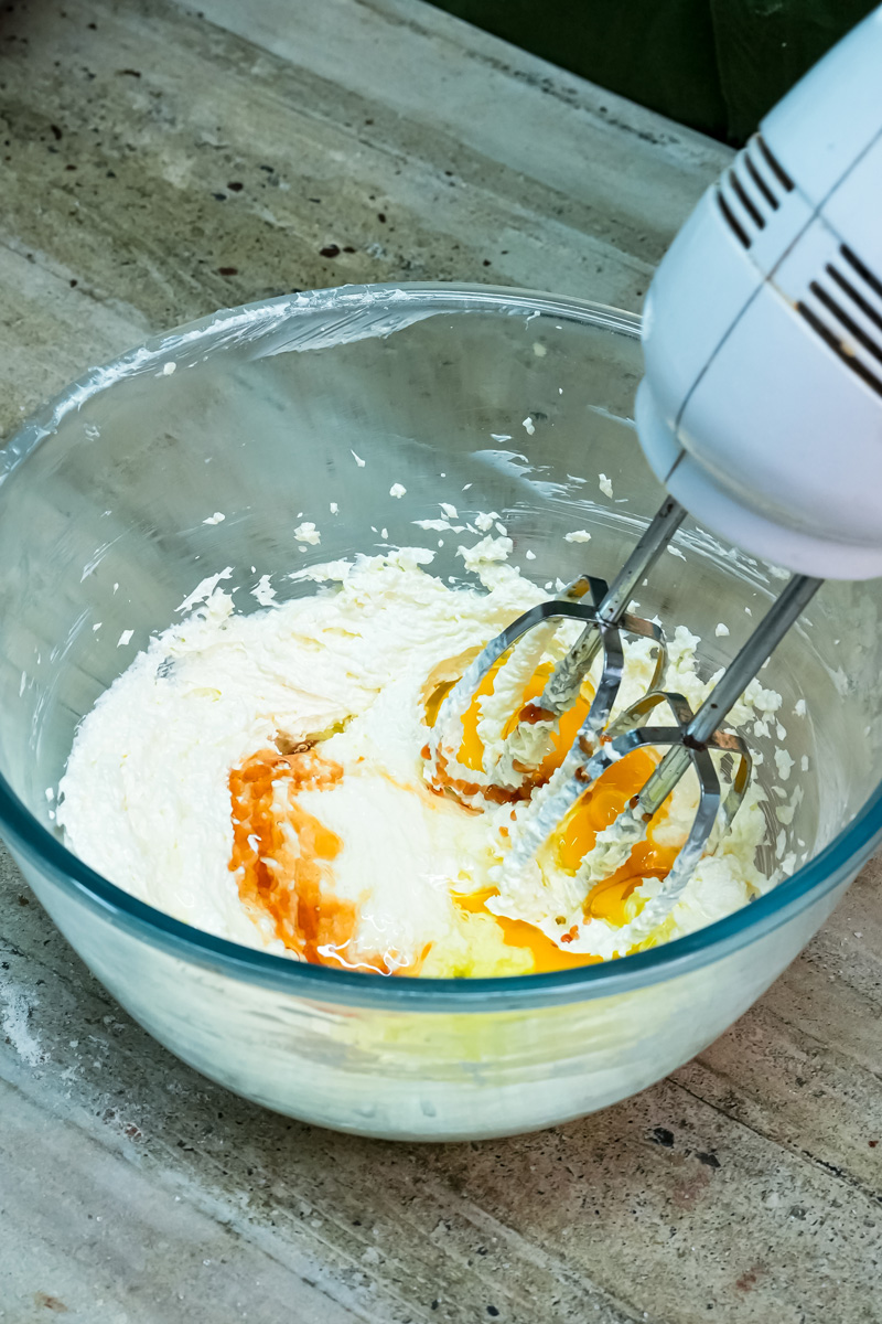 A hand mixer blending egg and vanilla extract into the creamed butter and sugar mixture for spritz cookies.