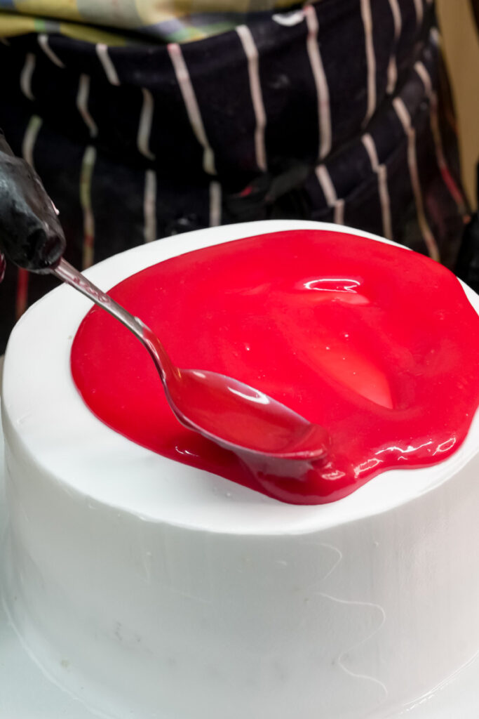 A baker spreading red mirror glaze on top of a cake with a spoon