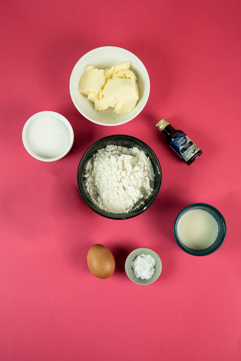 A top-down shot of spritz cookie ingredients on a pink background, including butter, flour, sugar, an egg, vanilla extract, salt, and milk