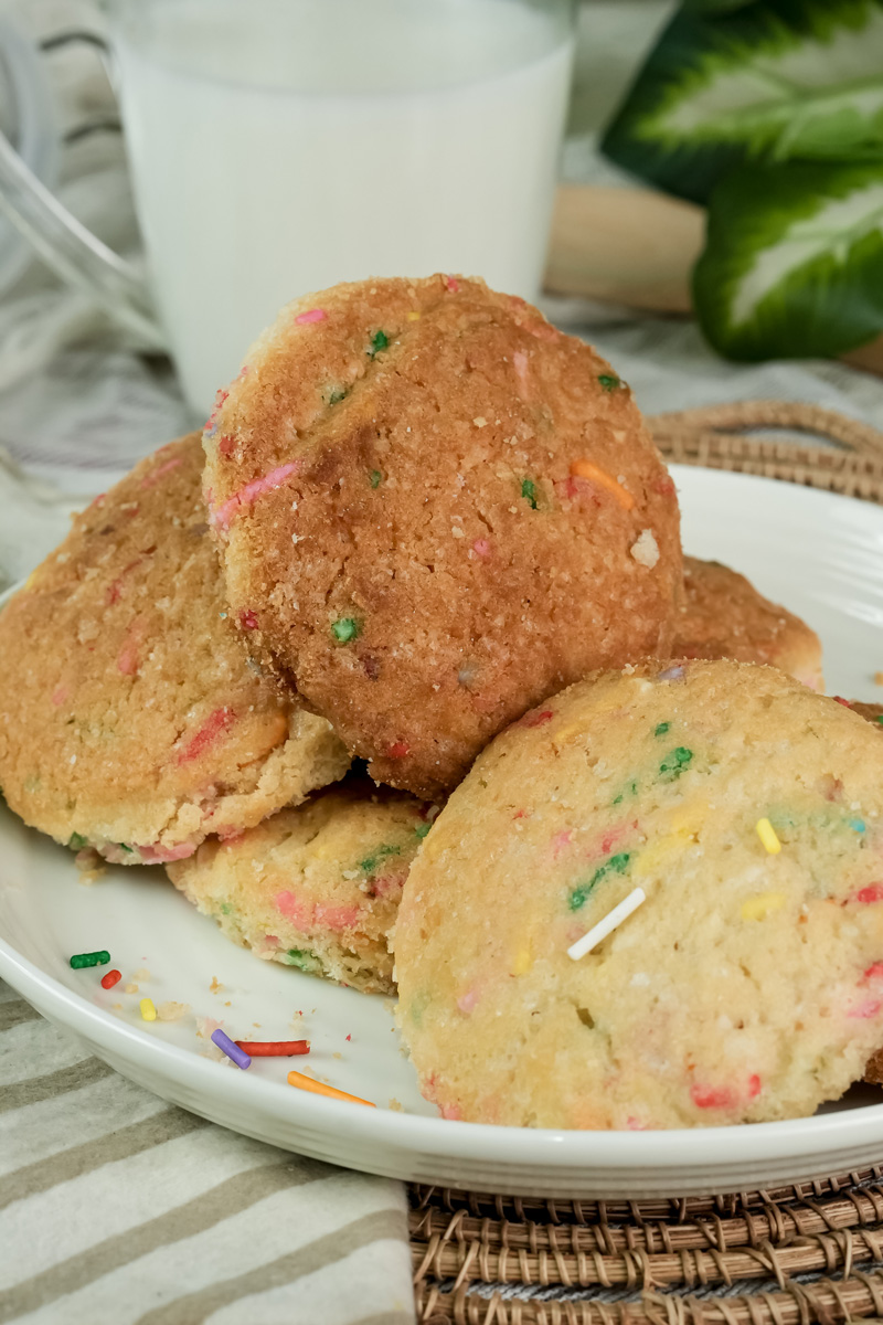 A stack of baked Funfetti cookies on a white plate with a glass of milk