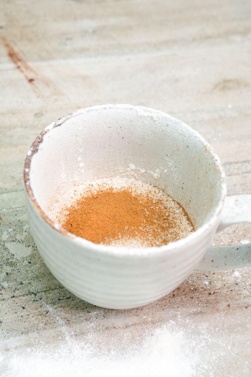 Mixing dry ingredients of flour, sugar, and cinnamon in a white ceramic mug
