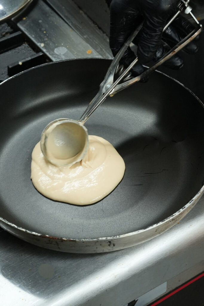 Using a portion scoop to pour batter into a non-stick frying pan